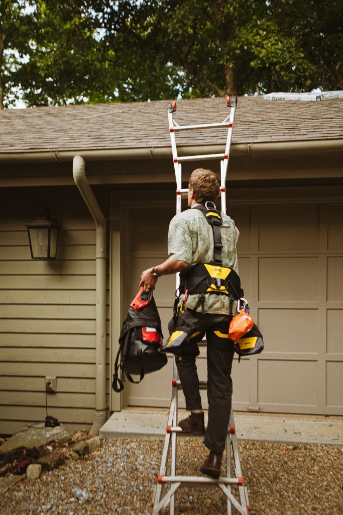 Roofer wearing a safety harness climbing an aluminium extension ladder set against a house